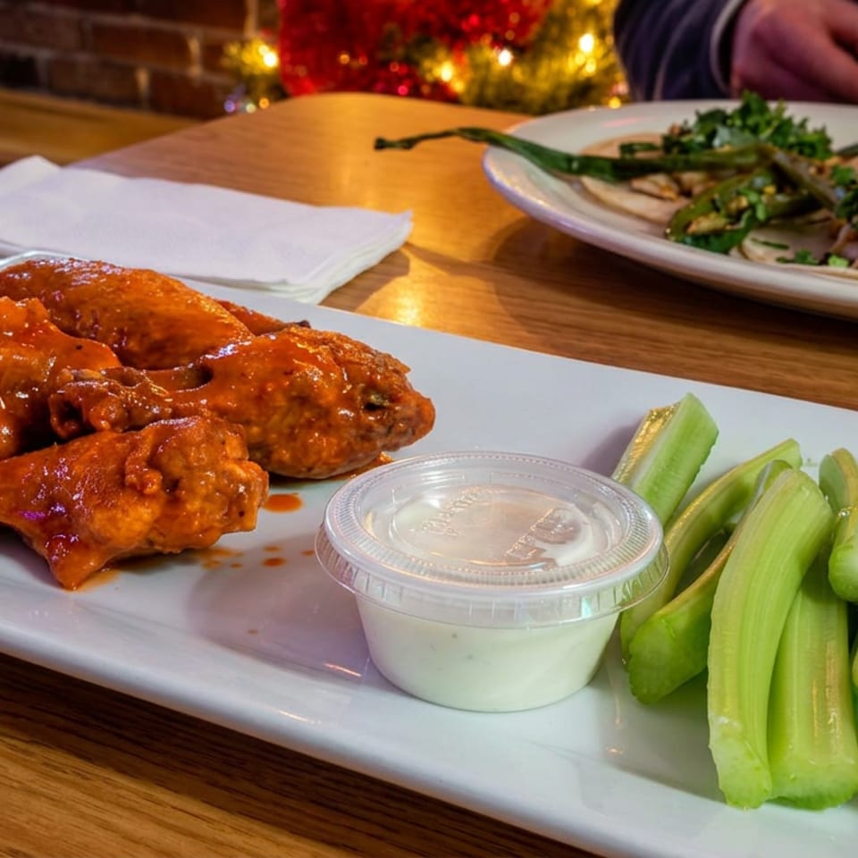 Buffalo chicken wings with celery sticks and ranch dipping sauce on a plate, alongside a garnished entrée in a cozy restaurant.