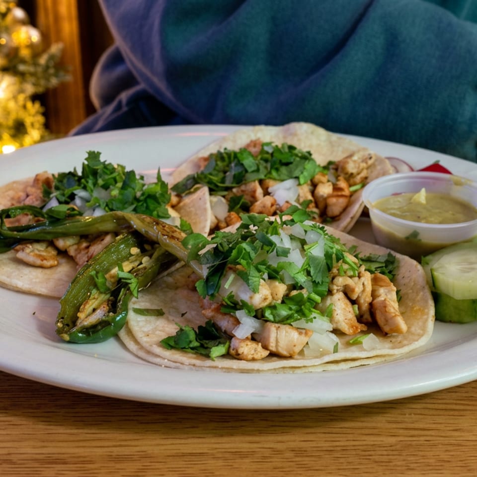 Plate of chicken tacos topped with cilantro and onions, grilled peppers, cucumber slices, and green salsa at a warmly lit restaurant.