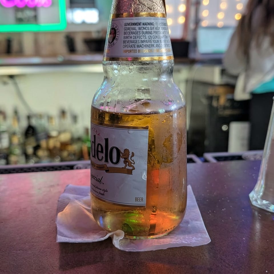 Open Modelo Especial beer bottle on a napkin beside a salt shaker at a neon-lit bar.