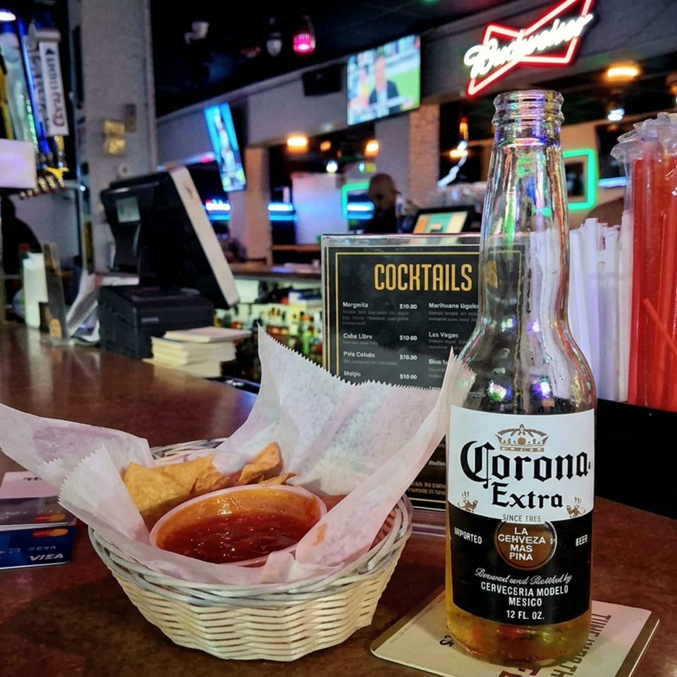 Bar counter with basket of tortilla chips and salsa, Corona beer, cocktail menu, TVs, and patrons in the background.